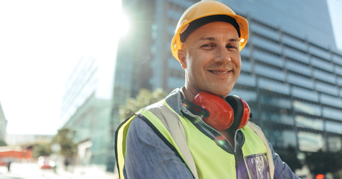 A worker wears a yellow hard hat, a high-visibility vest, and red noise-canceling headphones around their neck.