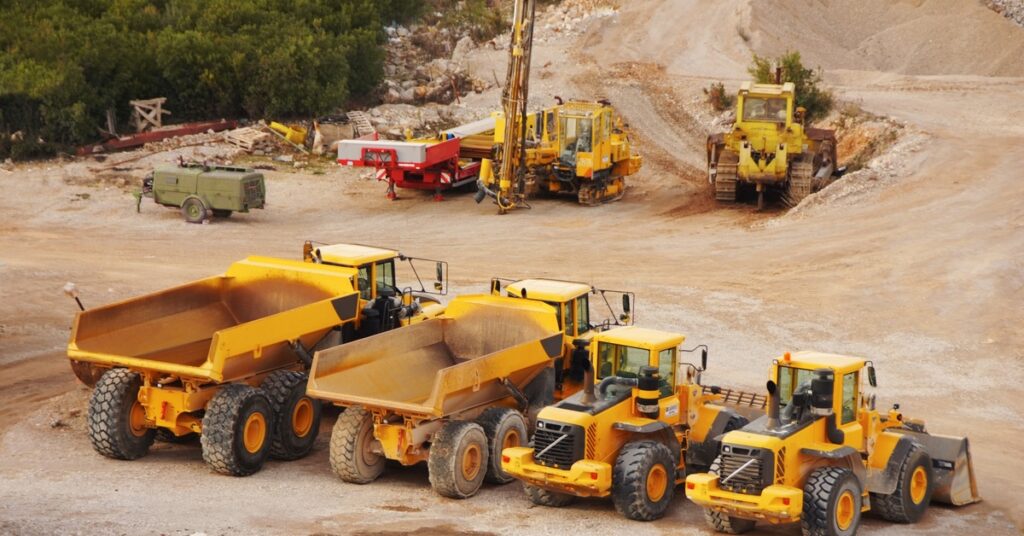 Multiple yellow mining vehicles sit next to each other in an empty mining quarry. There's a field of trees to the left.