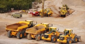 Multiple yellow mining vehicles sit next to each other in an empty mining quarry. There's a field of trees to the left.