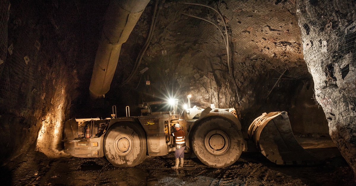 A large mining vehicle navigates a tunnel for a hard rock mine. Bright lights shine above the equipment.