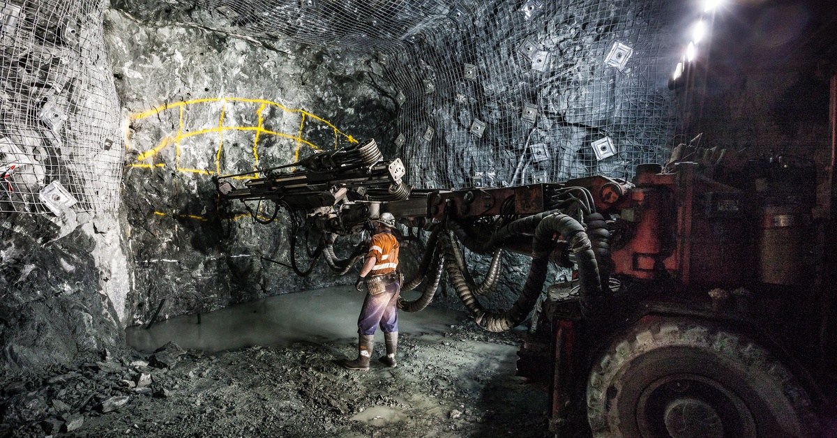A miner stands next to a large tunneling vehicle that is breaking apart rocks in a mine.