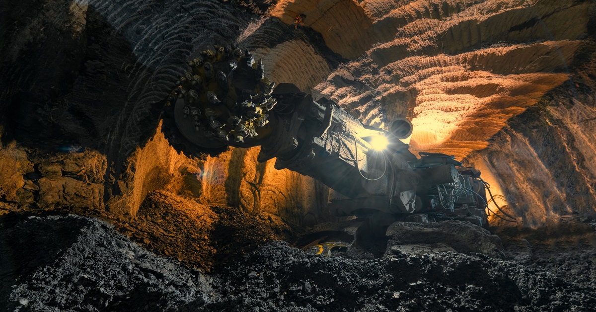 A large drilling machine operates in an underground mine, carving out the tunnel with its large metal drill.