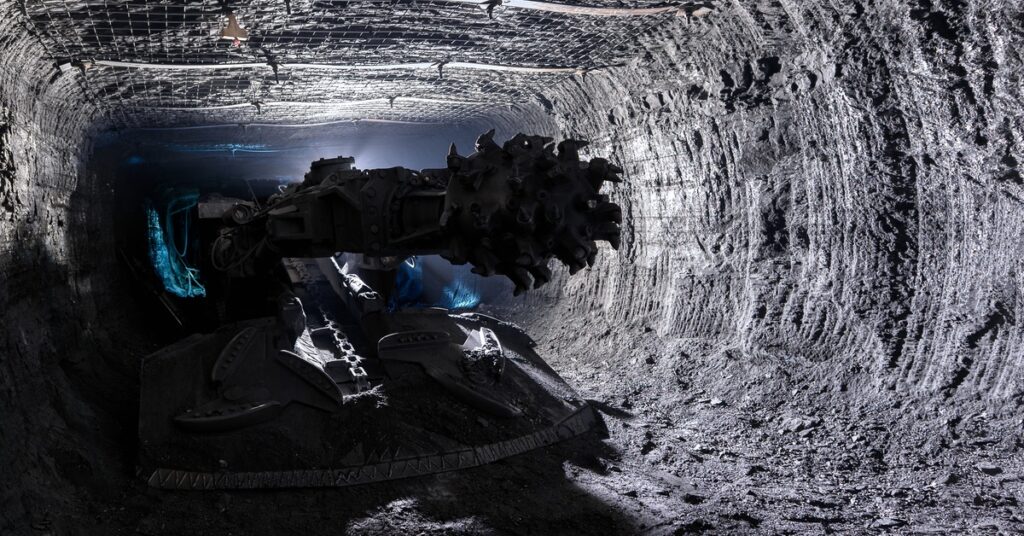 A shearer drill machine operates in an underground mine, with netting on the ceiling to prevent rocks from falling.
