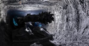 A shearer drill machine operates in an underground mine, with netting on the ceiling to prevent rocks from falling.