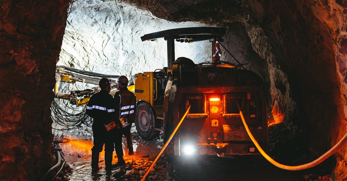 An underground mining machine navigates a mining tunnel with two workers in high-visibility vests next to it.