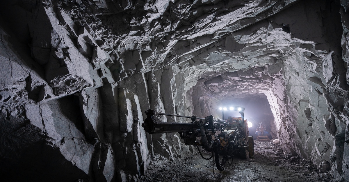 In the distance, there are multiple mining machines working in a large underground mine lit by a large light.