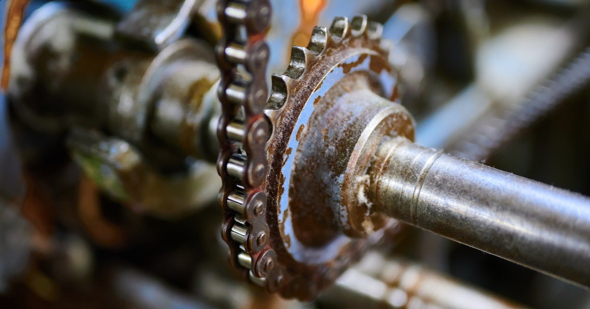 A machine with metal gear and chain at close range that features worn pieces, rust, and discoloration.