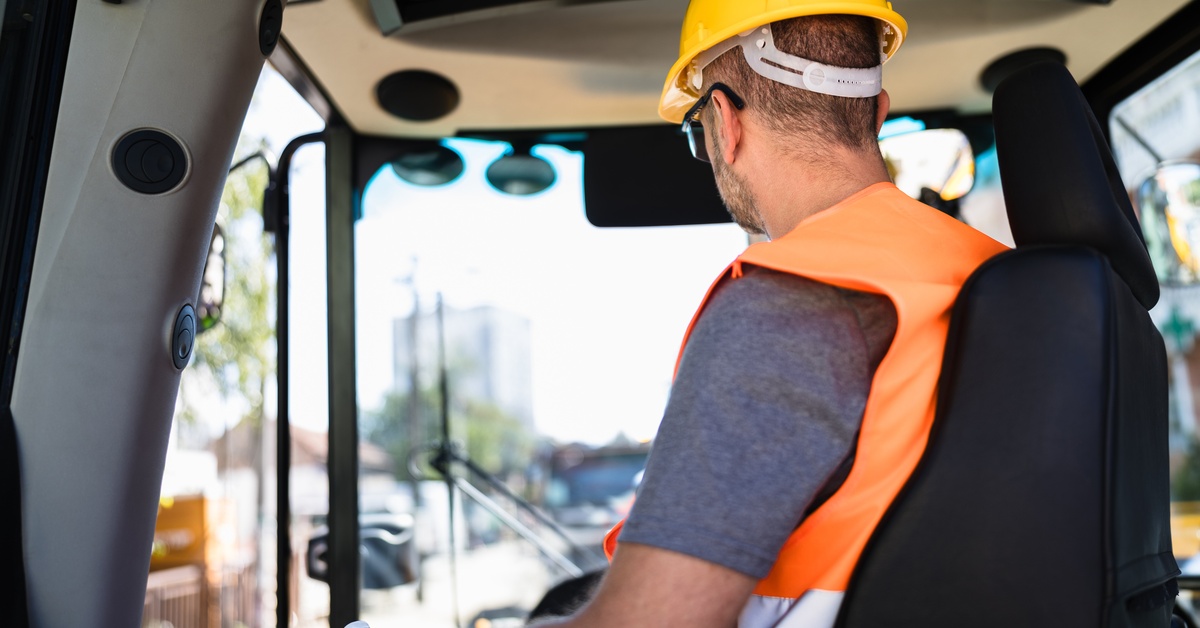 A worker in a hard hat and orange high-vis vest operates a piece of heavy machinery with a blurred city in the distance.