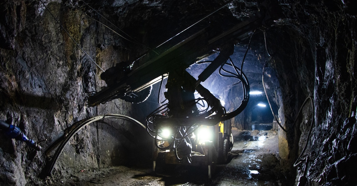 In a dark underground mine, a yellow vehicle operates in the distance. Lights on the vehicle and ceiling light the tunnel.