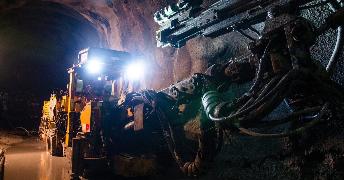 A yellow mining vehicle operates in a dark underground tunnel. Lights on top of the vehicle light up the tunnel.
