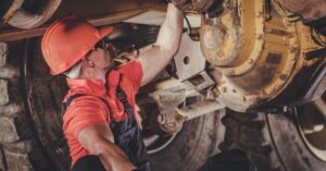 A worker sits on the ground wearing a hard hat, overalls, and gloves while they check the underside of a mining truck.