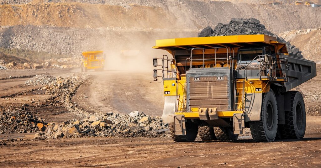 A large yellow mining vehicle drives through a mining site while hauling a full load of black material.