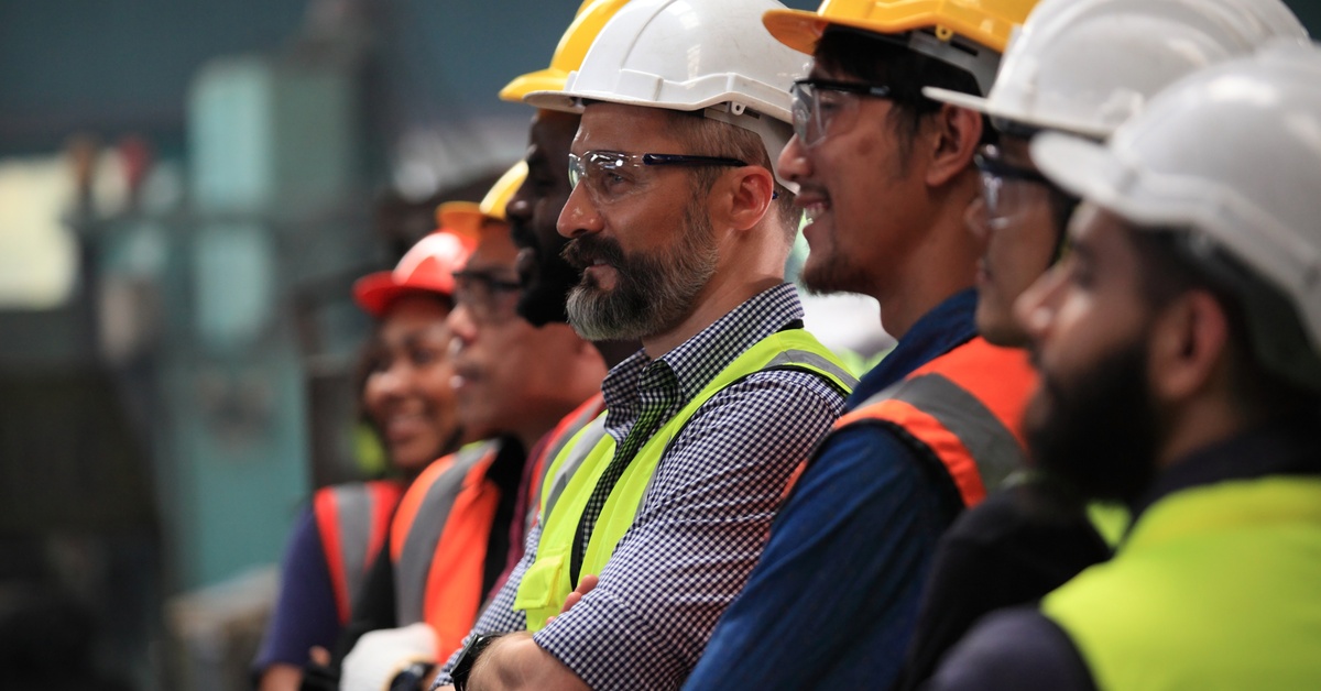 A group of people all wearing high-visibility vests and hard hats smile and look to the side. Some are wearing goggles.