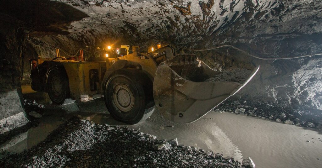 A large yellow load haul dumper machine drives through an underground mine with lights on top of it.