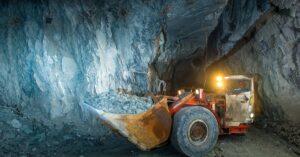 A small mining vehicle is in a dark tunnel, lighting up the tunnel with lights and holding a large amount of material.