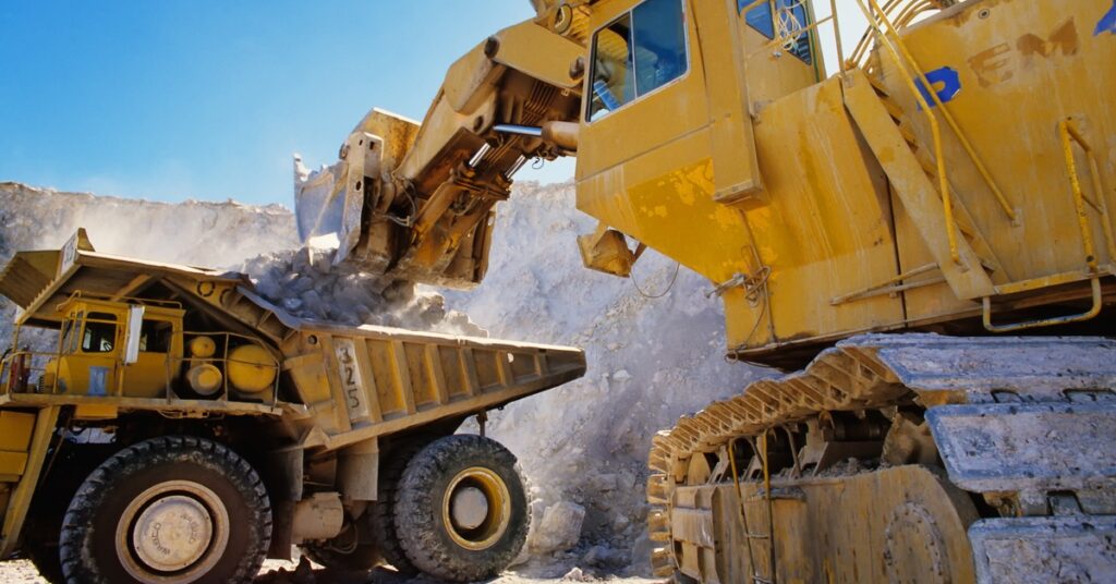 A large piece of mining equipment moves material into the back of a yellow truck at a mining site.