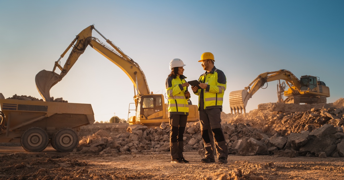 Two workers wearing hard hats and high-visibility vests stand at a mining site and talk to each other.