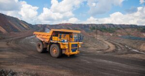 A large yellow mining dump truck drives around a mining quarry. There is a blue sky with white clouds in the distance behind the mining quarry.
