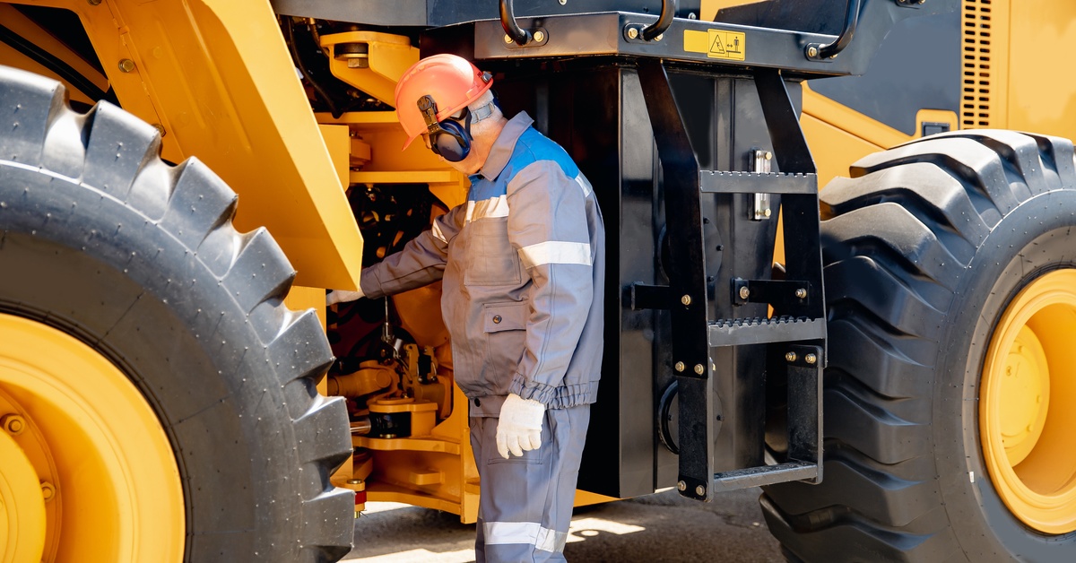 A mechanic wearing a work suit and a hard hat helmet examines the equipment in a large yellow mining vehicle.