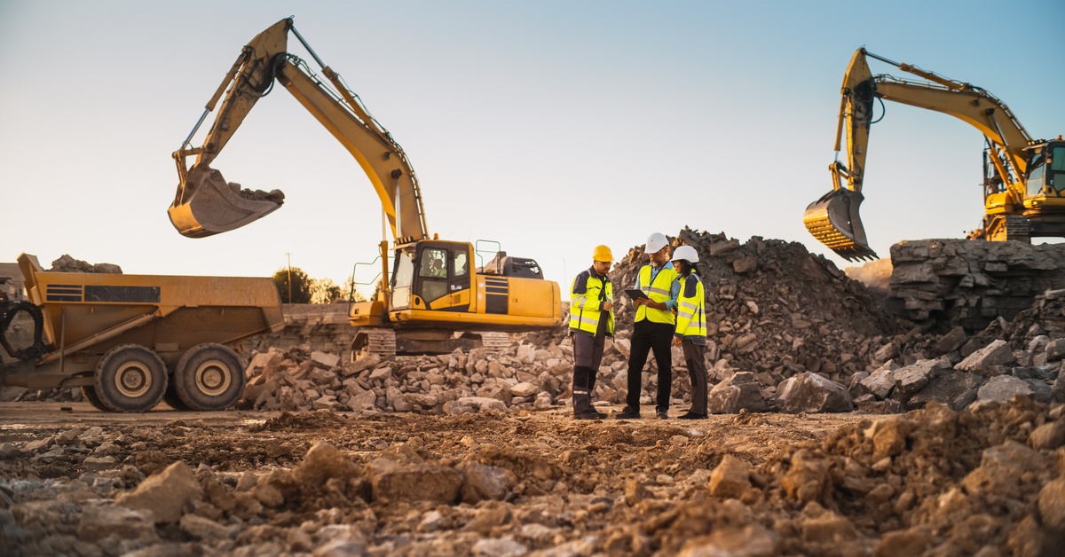 Three workers in vests and hard hats talk to each other in the middle of a mining site, surrounded by machines.