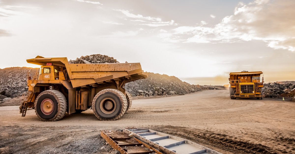 Two mining vehicles transporting minerals on a dirt road. There are large mounds of rocks and gravel in the background.