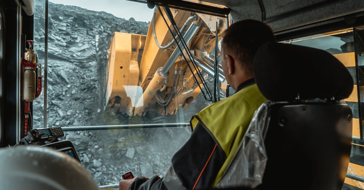 A man in a yellow vest operates a mining vehicle to dig through a tall mound of rocky gray material.