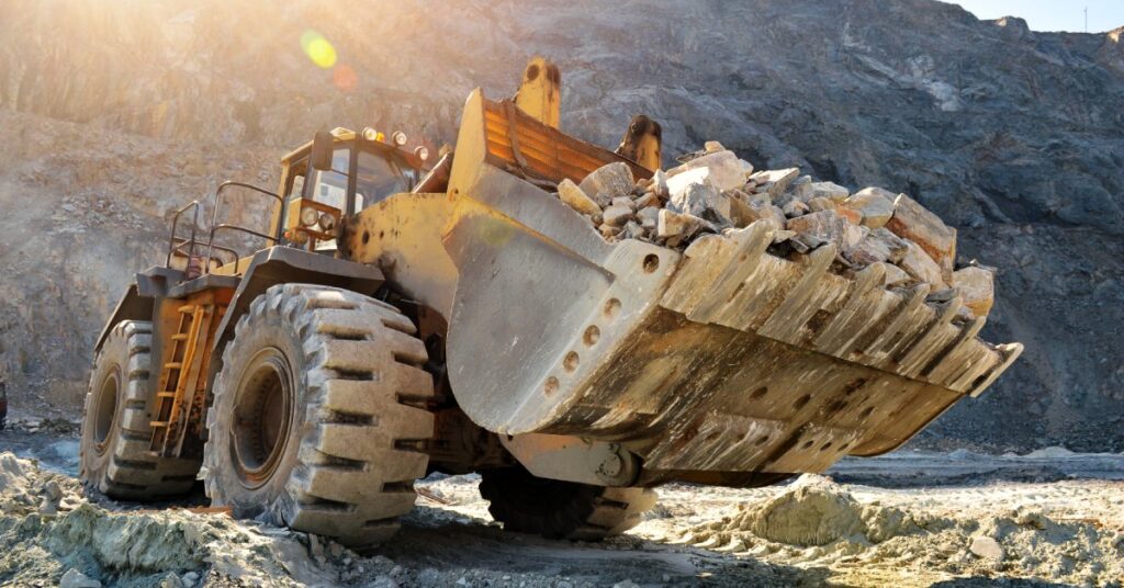 A mining vehicle parked at the bottom of a trench. The vehicle's bucket holds large rocks and rubble.
