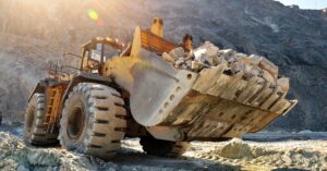 A mining vehicle parked at the bottom of a trench. The vehicle's bucket holds large rocks and rubble.
