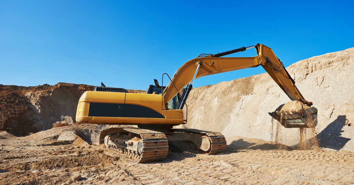 An excavator shoveling sand at a dig site. There are tread marks in the sand from the vehicle’s track pad.