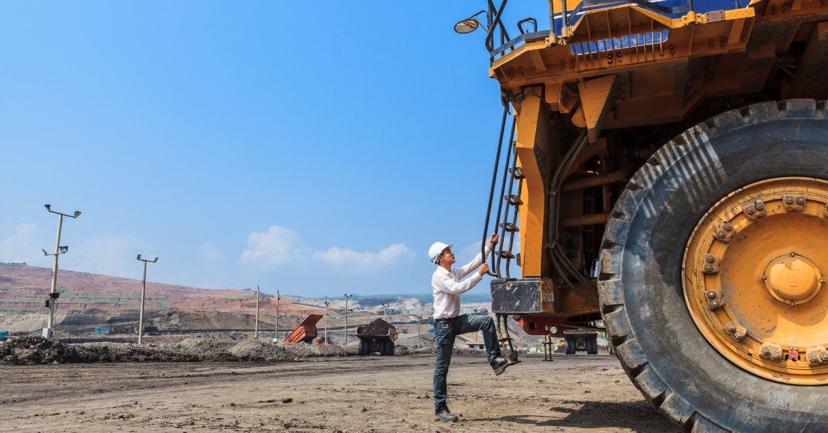 A person climbing the ladder of a large mining vehicle. They are wearing a white hard hat and a long-sleeved shirt.