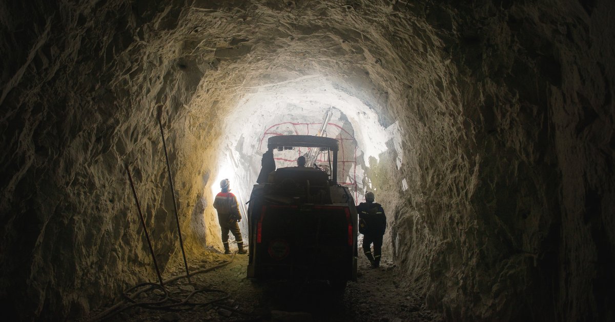 Three mining operators exiting a dark mine. One of the operators is driving a large hard rock mining vehicle.