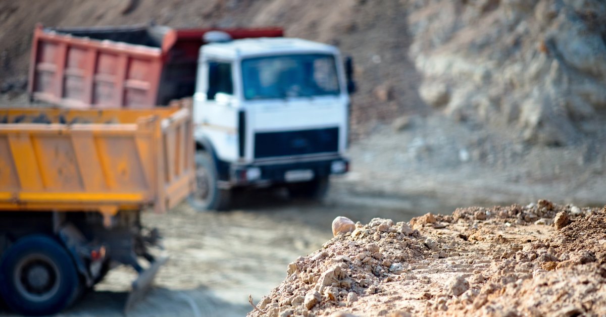 Two heavy mining trucks with large buckets parked at a dig site. There is a pile of gravel and rocks in the foreground.