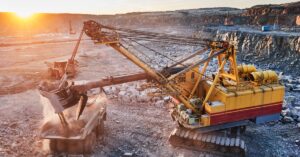 A mining excavator loading gravel and minerals into a dump truck at a mining site. There are large rocks and rubble nearby.