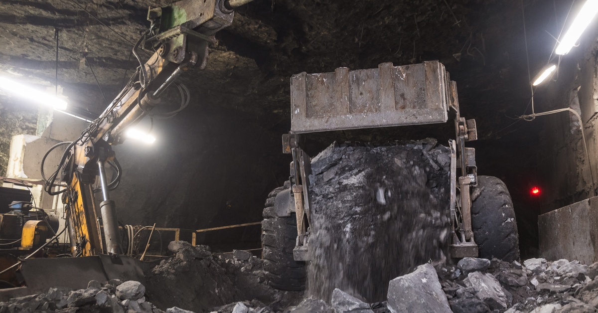 Two mining vehicles parked in an underground mine. One of the vehicles is dumping black gravel onto the ground.
