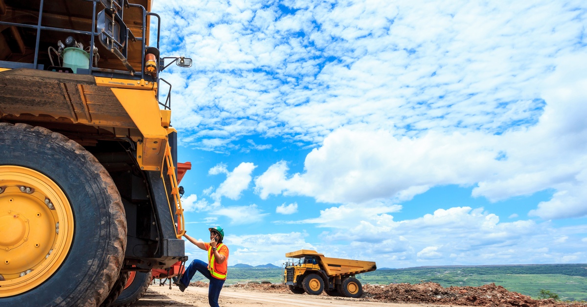 A person climbing the ladder of a mining vehicle with a walkie talkie in their hand. They are wearing a reflective vest.