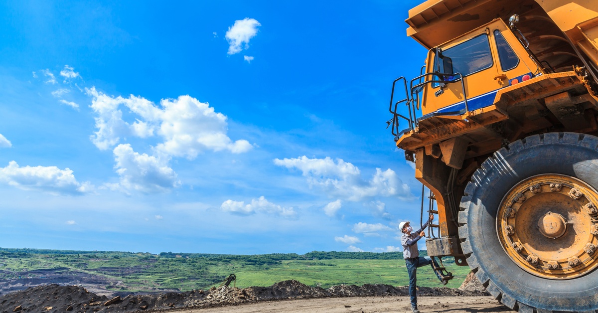 A mining operator climbing the ladder of a large off-highway vehicle. The man is wearing a white hard hat and a mask.