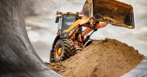 A mining vehicle sitting on top of a mound of dirt. Attached to the vehicle is a large bucket tilted forward.
