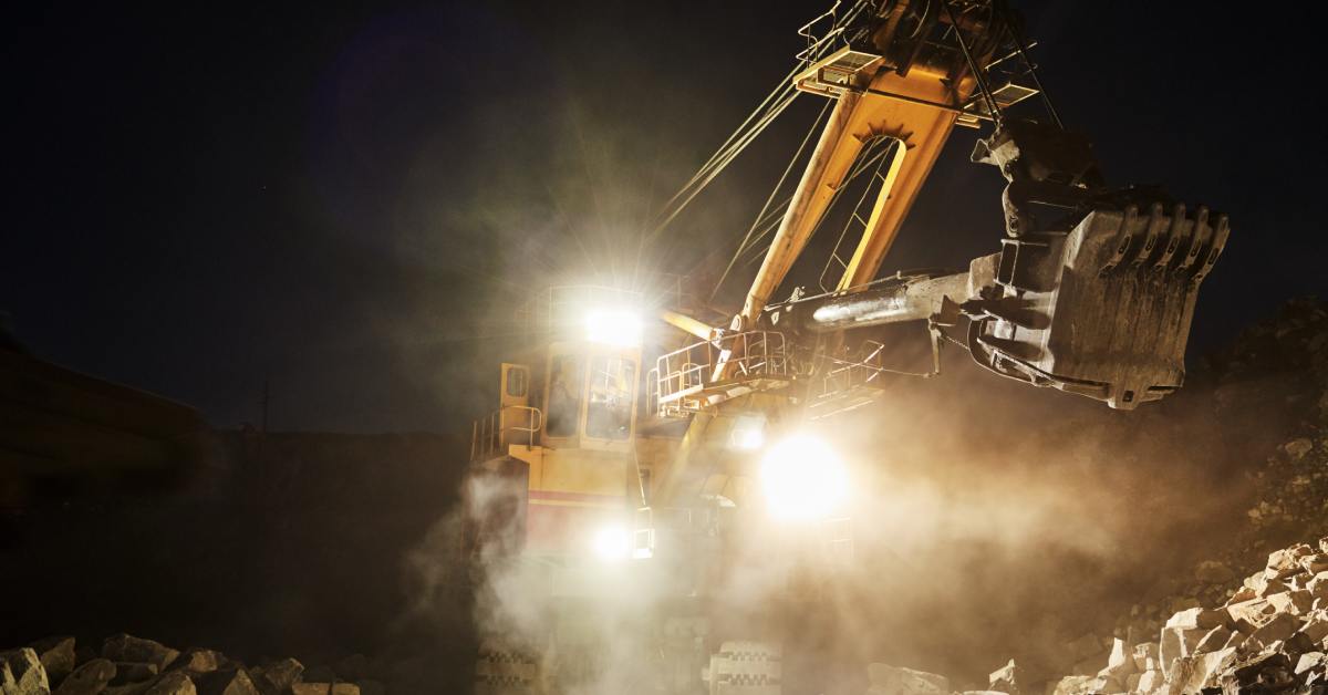 An excavator shoveling minerals and gravel in a dark quarry. There are bright lights at the front of the vehicle.