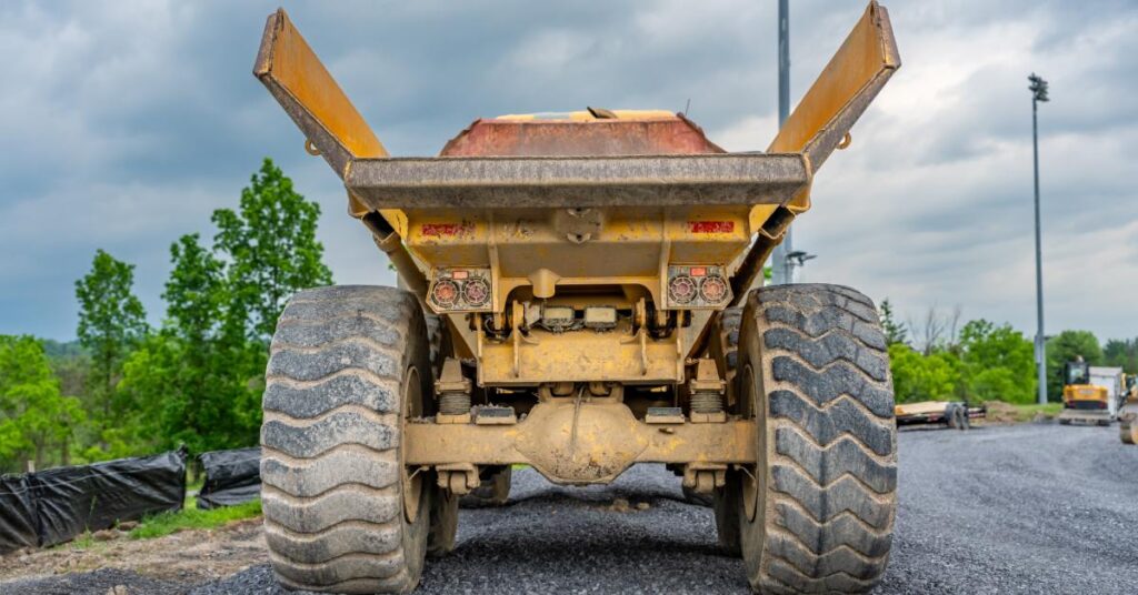 The backside of a large dump truck on a gravel road. The bed of the truck is empty, and dirt covers the wheels.