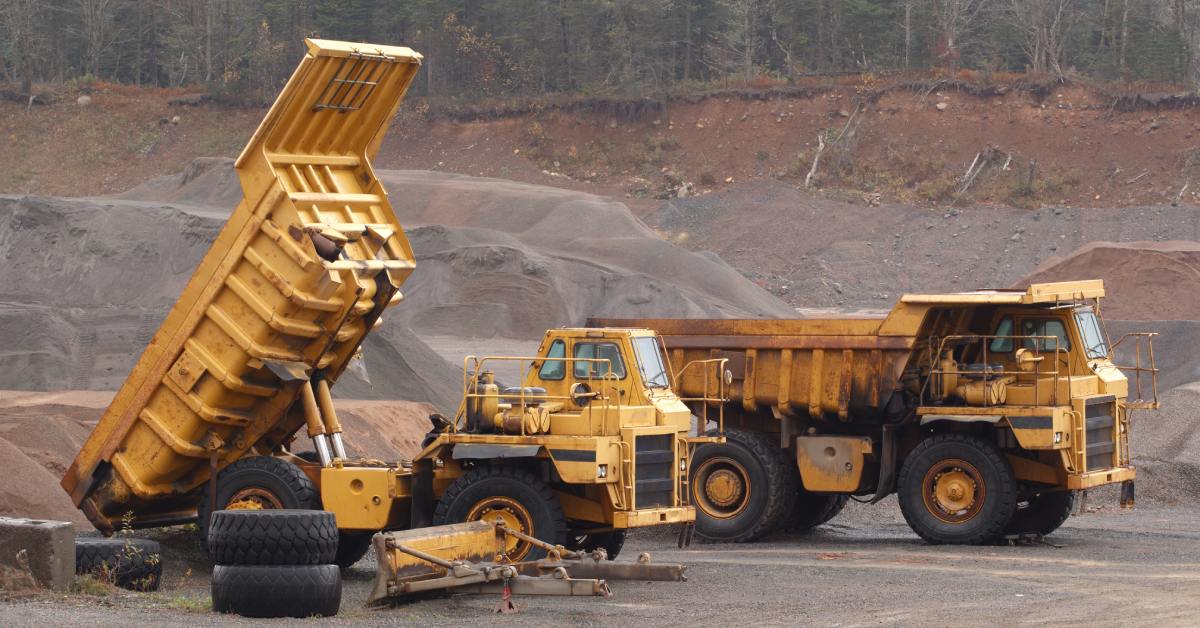 Two yellow off-highway vehicles are parked in a pit with mounds of dirt surrounding them. One truck bed is lifted in the air.