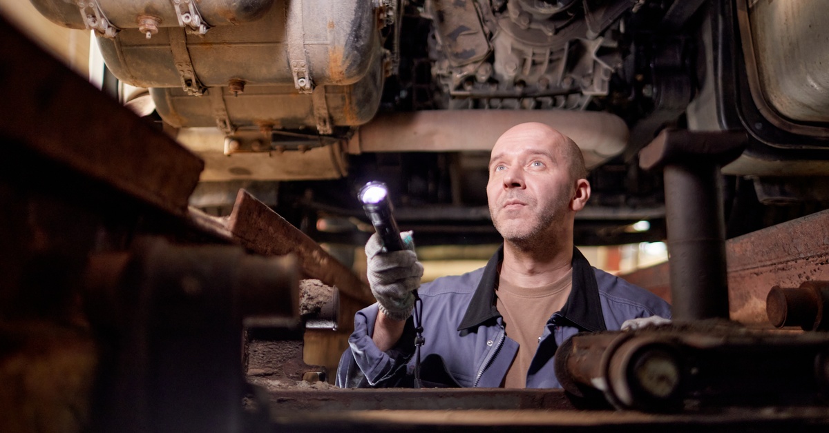 A person wearing blue coveralls holds a flashlight and looks at the undercarriage of heavy machinery.