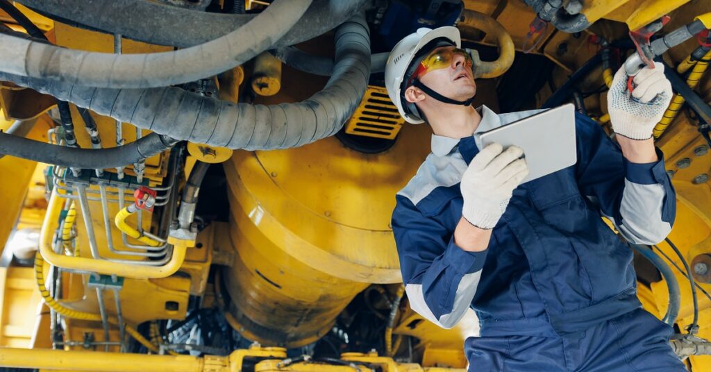 A man wearing a hard hat, safety goggles, and coveralls holds a tablet and examines the undercarriage of heavy machinery.