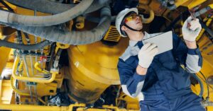 A man wearing a hard hat, safety goggles, and coveralls holds a tablet and examines the undercarriage of heavy machinery.