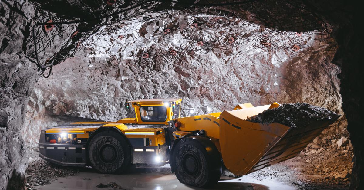 A yellow mining loader with a full bucket in a dark, rocky tunnel. Bright lights reflect on wet, muddy ground.