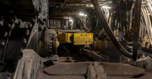 A dark, dusty underground mine with heavy machinery, a yellow metal component, and industrial cables under harsh lighting.