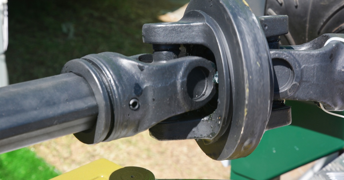 A close-up of a universal joint with dark metal yokes, a greased center, and shafts against a blurred outdoor background.