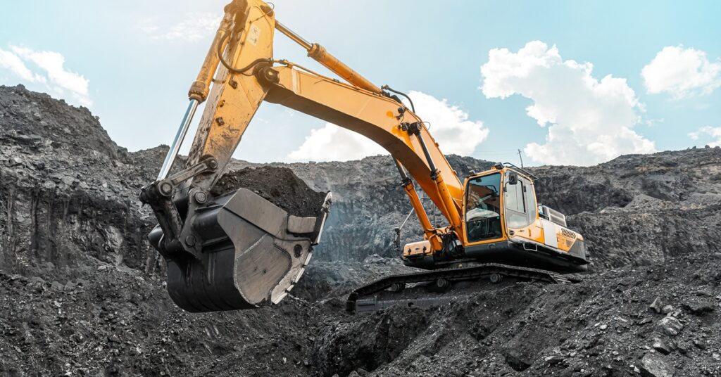 A yellow excavator is positioned deep in a quarry. The claw has a heaping pile of black coal and draws it upward.