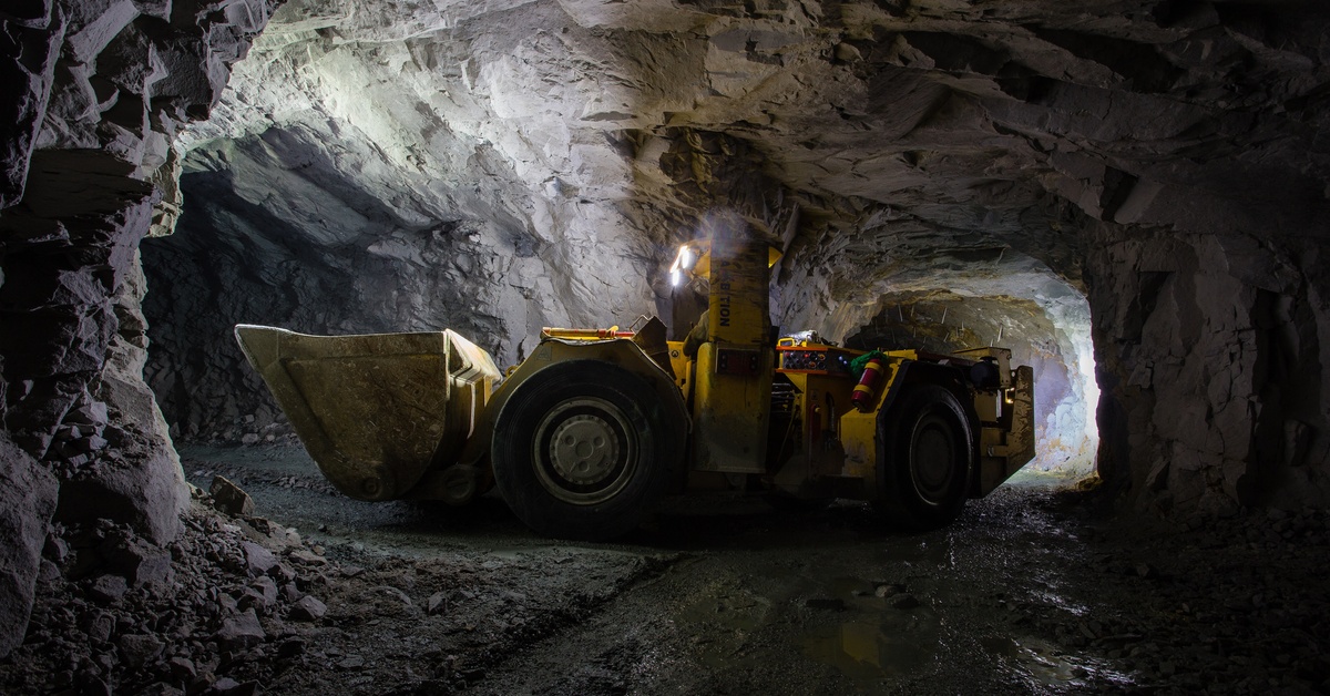 A yellow mining loader with an extended bucket extended. It sits in a dark underground tunnel illuminated by vehicle lights.