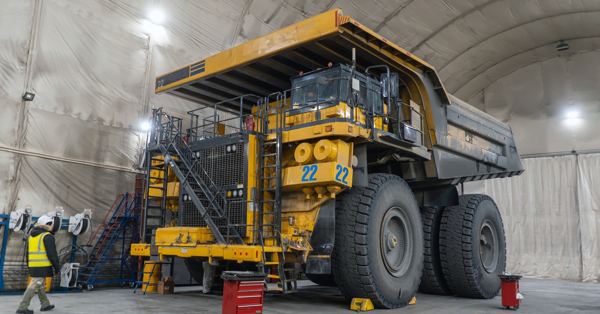 A yellow mining dump truck is parked in a large repair warehouse. A mechanic walks toward the machinery.