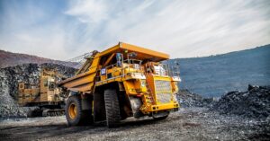 A yellow mining truck is driving up along a quarry with piles of rock surrounding the jobsite. The sky is clear and blue.